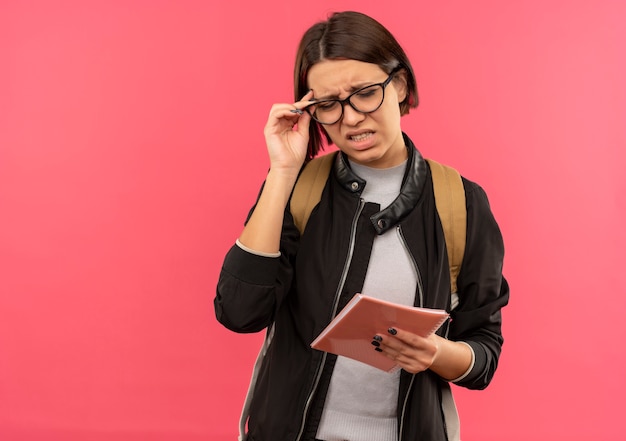 Menina jovem estudante irritada usando óculos e uma bolsa com as costas segurando um bloco de notas e seus óculos com os olhos fechados, isolado em rosa com espaço de cópia