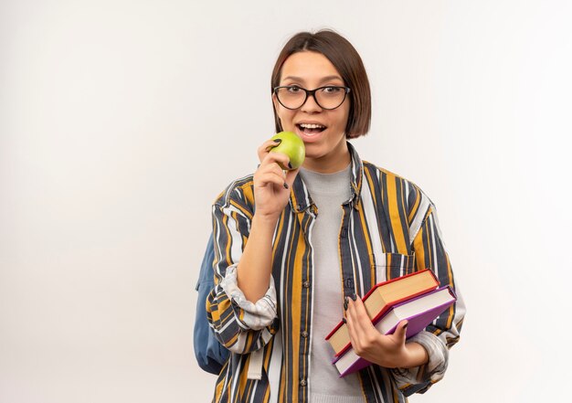 Menina jovem estudante usando óculos e mochila segurando livros e se preparando para morder uma maçã isolada no fundo branco com espaço de cópia