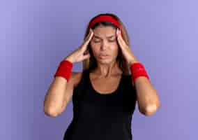 Foto grátis menina jovem fitness em roupa esportiva preta e bandana vermelha, parecendo doente, tocando sua cabeça com as mãos, sofrendo de forte dor de cabeça, em pé sobre a parede azul