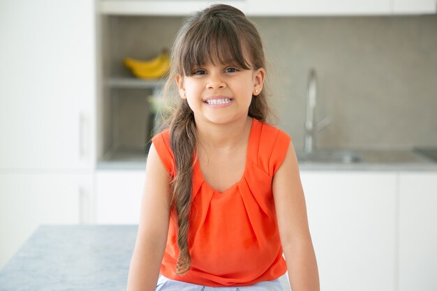 Menina latina alegre, de cabelos escuros, com camisa vermelha sem mangas, posando na cozinha