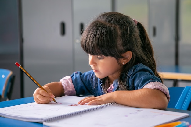 Foto grátis menina latina de cabelos pensativos sentada na carteira da escola e desenhando em seu caderno