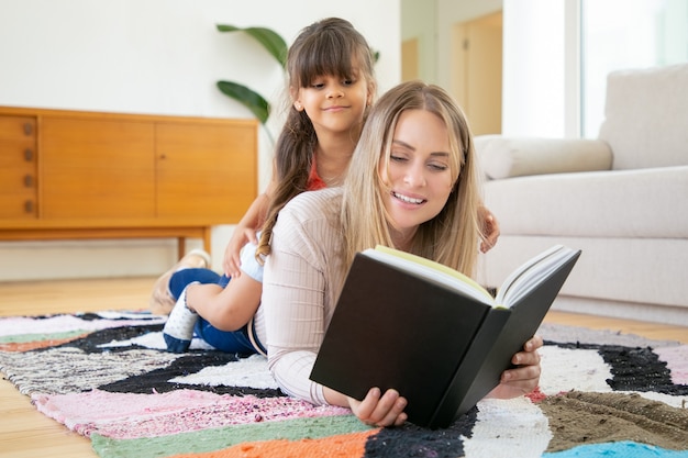 Menina latina sentada nas costas da mãe, sorrindo e olhando para o livro.