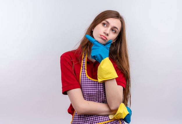 Foto grátis menina linda com avental e luvas de borracha olhando para cima com a mão no queixo e expressão pensativa