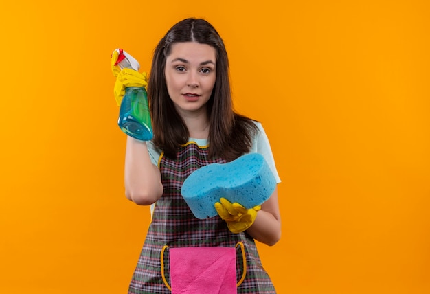 Menina linda com avental e luvas de borracha segurando spray de limpeza e esponja olhando para a câmera sorrindo confiante