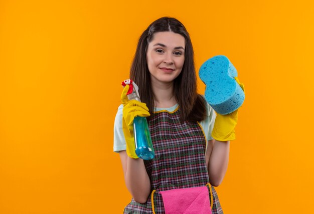 Menina linda com avental e luvas de borracha segurando spray de limpeza e esponja olhando para a câmera sorrindo confiante