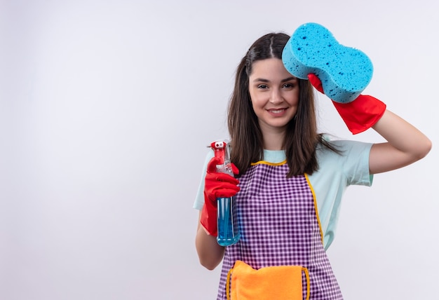 Menina linda com avental e luvas de borracha segurando spray de limpeza e mostrando a esponja sorrindo alegremente