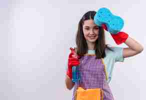 Foto grátis menina linda com avental e luvas de borracha segurando spray de limpeza e mostrando a esponja sorrindo alegremente