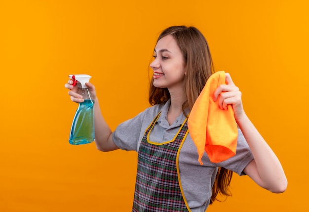 Menina linda com avental segurando spray de limpeza e tapete olhando para o lado sorrindo, pronta para limpar