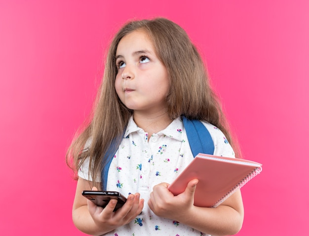 Menina linda com cabelo comprido com mochila segurando notebook e smartphone olhando para cima perplexa em pé na rosa