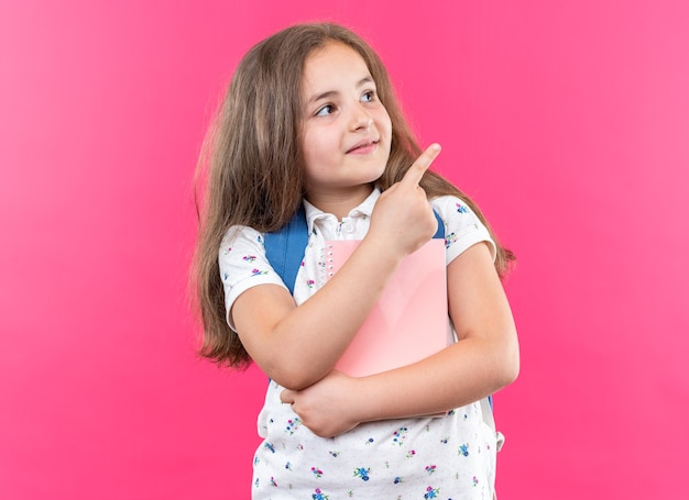 Menina linda com cabelo comprido com mochila segurando o caderno olhando para cima com um sorriso no rosto apontando com o dedo indicador para o lado em pé sobre a parede rosa