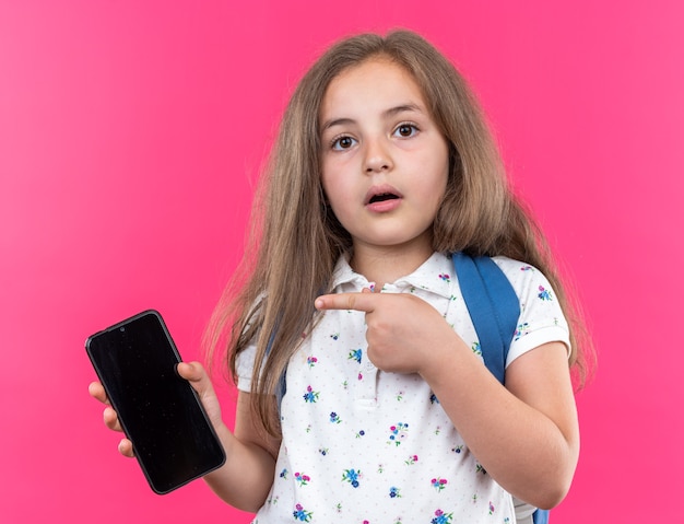 Menina linda com cabelo comprido com mochila segurando o smartphone apontando com o dedo indicador para ele surpresa em pé na rosa