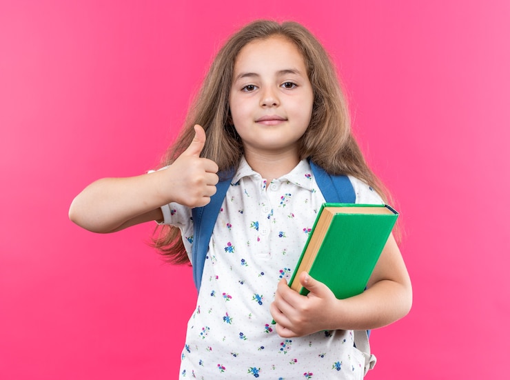 Foto grátis menina linda com cabelo comprido com mochila segurando um caderno olhando para frente sorrindo confiante mostrando os polegares em pé sobre a parede rosa