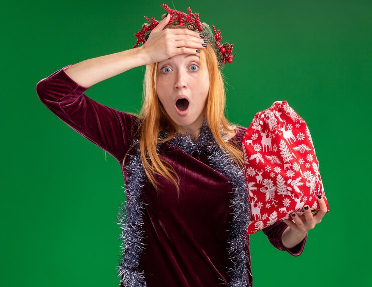 Foto grátis menina linda com medo de vestido vermelho com coroa de flores e guirlanda no pescoço segurando uma sacola de natal colocando a mão na testa isolada na parede verde