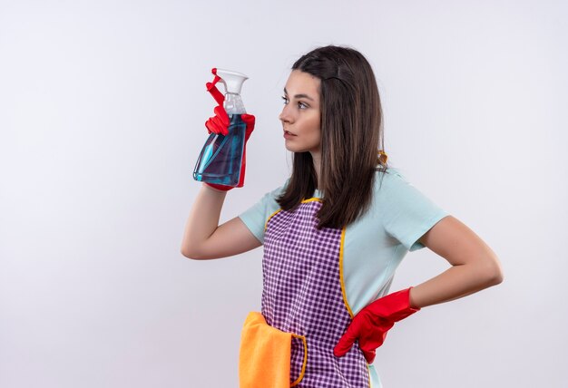 Menina linda de avental e luvas de borracha segurando spray de limpeza e olhando para o lado com uma cara séria