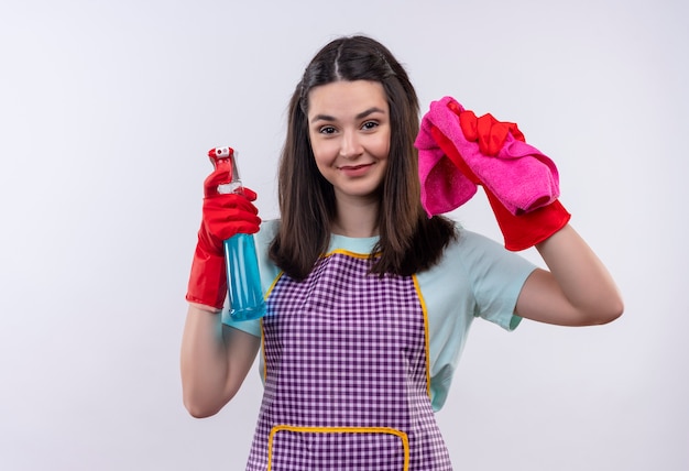 Menina linda de avental e luvas de borracha segurando spray de limpeza e tapete, olhando para a câmera, sorrindo alegremente, pronta para limpar