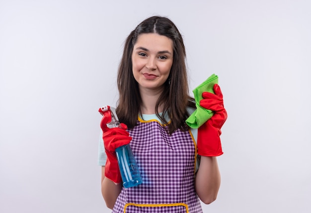 Menina linda de avental e luvas de borracha segurando spray de limpeza e tapete sorrindo confiante, pronta para limpar