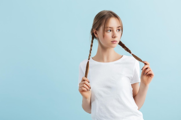 Menina linda de camiseta branca segurando duas tranças de cabelo nas mãos pensativamente olhando na câmera sobre fundo azul isolado