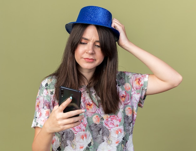 Menina linda e desagradável com chapéu de festa segurando e olhando para o telefone, colocando a mão na cabeça