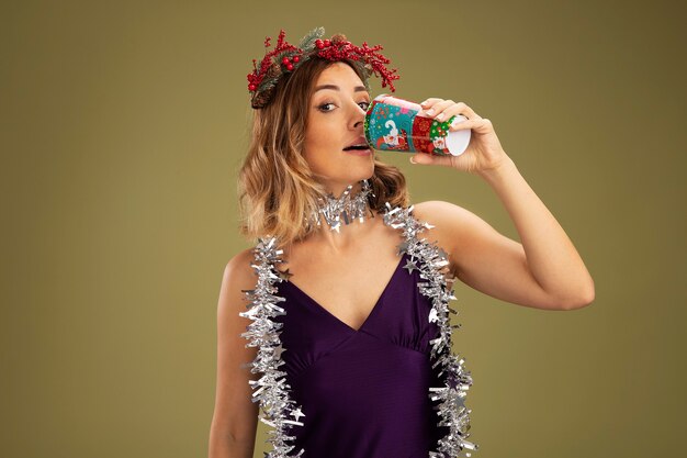 Menina linda e satisfeita com vestido roxo e grinalda com guirlanda no pescoço bebidas isoladas em fundo verde oliva