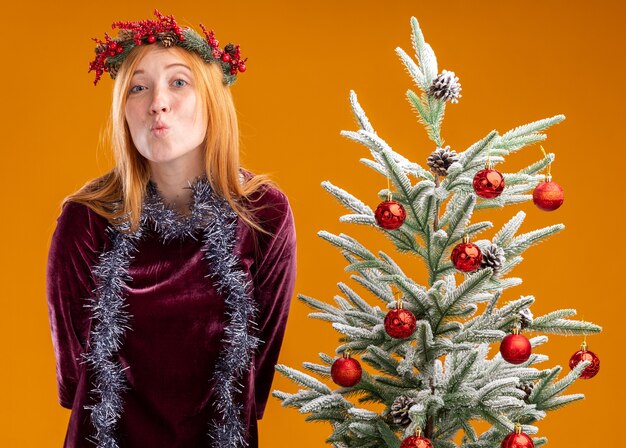 Menina linda e satisfeita em pé perto da árvore de Natal com vestido vermelho e grinalda com guirlanda no pescoço, mostrando gesto de beijo isolado em fundo laranja