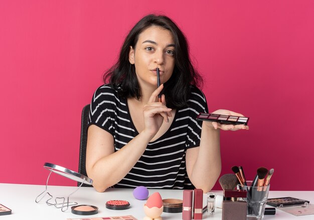 Menina linda e satisfeita se senta à mesa com ferramentas de maquiagem, aplicando sombra com pincel de maquiagem e colocando o pincel nos lábios isolados na parede rosa