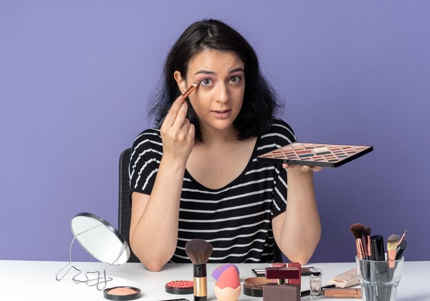 Menina linda e satisfeita sentada à mesa com ferramentas de maquiagem, aplicando a sombra com pincel de maquiagem isolado na parede azul