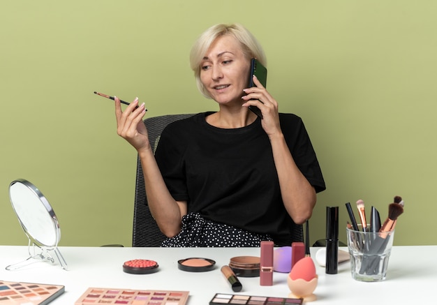 Foto grátis menina linda e satisfeita sentada à mesa com ferramentas de maquiagem falando ao telefone segurando o pincel de maquiagem isolado na parede verde oliva