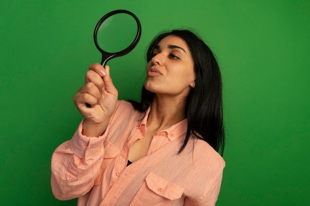 Menina linda e satisfeita vestindo camiseta rosa levantando e olhando para a lupa isolada na parede verde