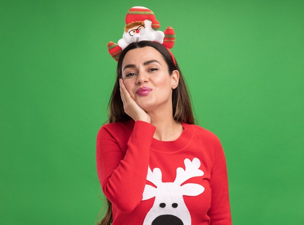 Menina linda e satisfeita vestindo um suéter de natal com uma argola de cabelo natalina colocando a mão na bochecha isolada sobre fundo verde