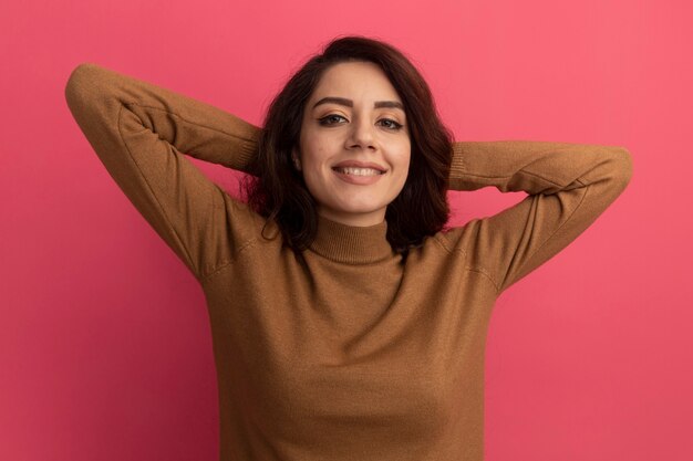 Menina linda e satisfeita vestindo uma blusa de gola alta, segurando as mãos atrás da cabeça, isolada na parede rosa