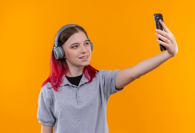 Menina linda e satisfeita vestindo uma camiseta cinza com fones de ouvido, tirando uma selfie em fundo amarelo isolado