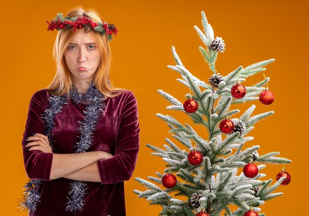 Menina linda e triste em pé perto da árvore de Natal com vestido vermelho e grinalda com guirlanda no pescoço, cruzando as mãos isoladas em um fundo laranja