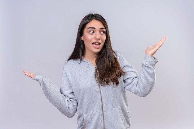 Menina linda em um capuz cinza olhando para longe com emoção chocada no rosto, parecendo incerta e confusa, sem resposta, espalhando as palmas das mãos sobre um fundo branco