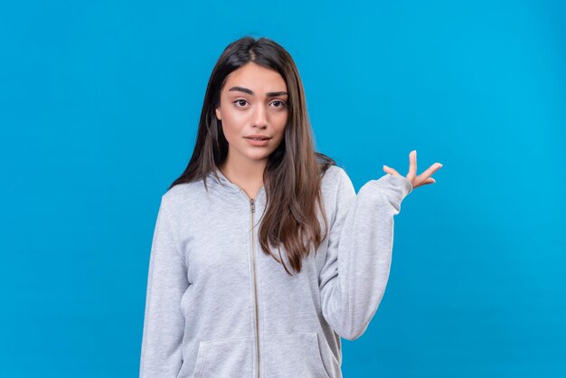 Menina linda em um casaco cinza com capuz olhando para a câmera com uma expressão instável em pé sobre um fundo azul