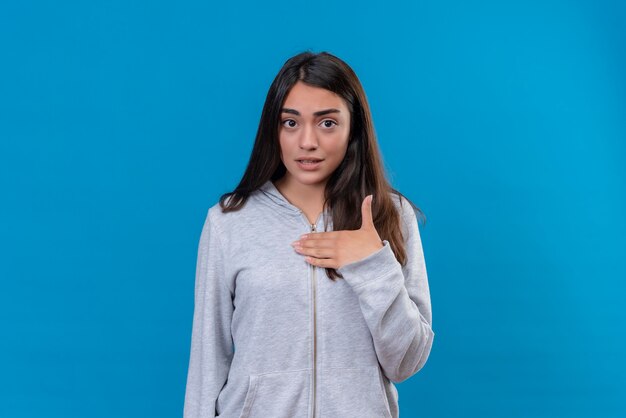 Menina linda em um casaco cinza com capuz olhando para a câmera, expressão insatisfeita no rosto, tocando-se em pé sobre um fundo azul