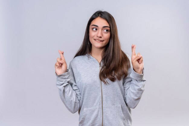 Menina linda em um casaco cinza com capuz olhando para longe e mordendo os lábios fazendo um gesto de boa sorte em pé sobre um fundo branco