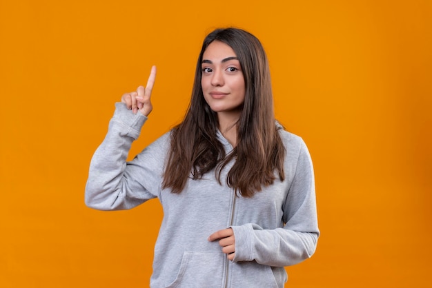 Menina linda em um casaco com capuz cinza apontando para cima tendo uma ótima ideia, sorrindo confiante em pé sobre um fundo laranja