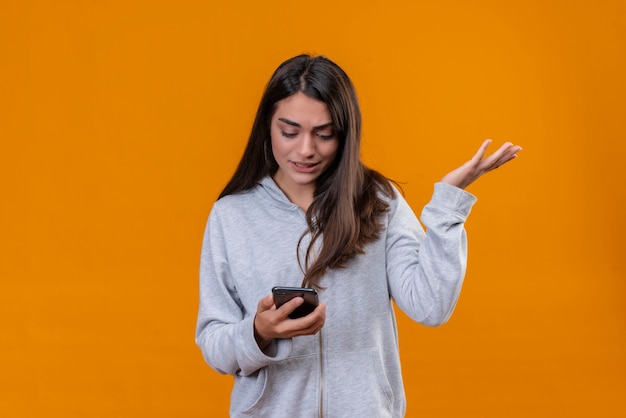 Menina linda em um casaco com capuz cinza com um telefone na mão olhando para o telefone com uma emoção de confusão em pé sobre um fundo laranja