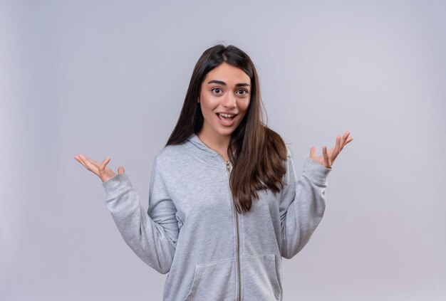 Menina linda em um casaco com capuz cinza olhando para a câmera com um sorriso no rosto, sem resposta, espalhando as palmas das mãos em pé sobre um fundo branco