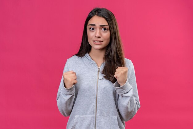 Menina linda em um casaco com capuz cinza olhando para a câmera com uma cara ansiosa preocupada em pé sobre um fundo rosa