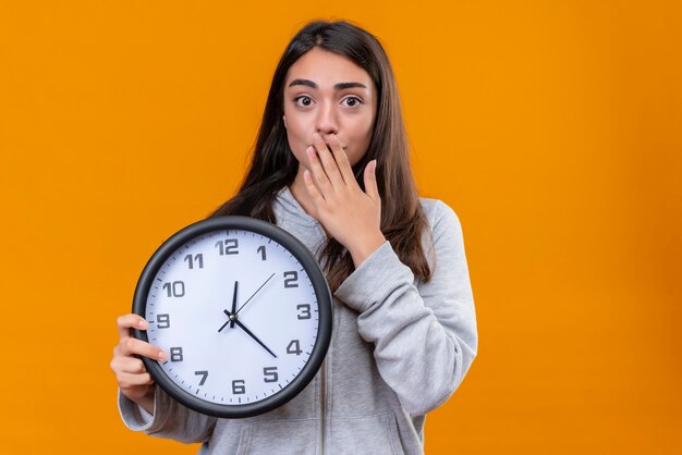 Menina linda em um casaco com capuz cinza segurando o relógio e olhando para a câmera com surpresa no rosto em pé sobre um fundo laranja