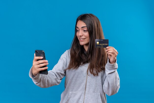 Menina linda em um casaco com capuz cinza segurando o telefone e mostrando o cartão de crédito para o telefone.