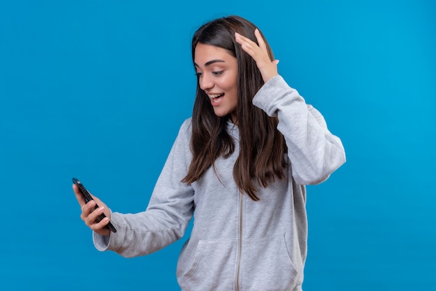 Foto grátis menina linda em um casaco com capuz cinza segurando o telefone e olhando para o telefone com um sorriso no rosto em pé sobre um fundo azul