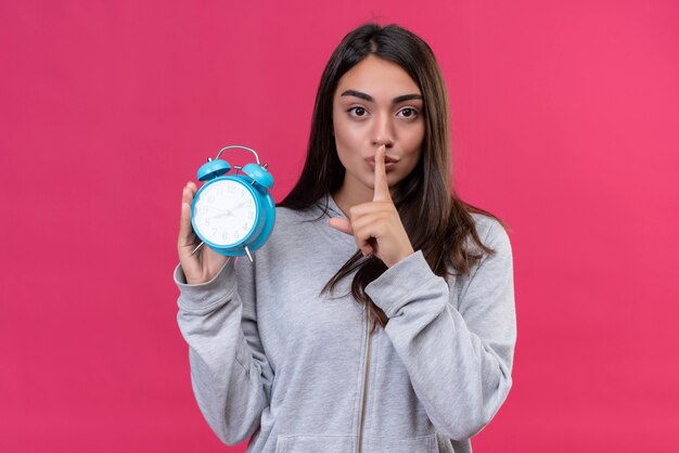 Menina linda em um casaco com capuz cinza segurando um relógio olhando para a câmera e fazendo um gesto de silêncio em pé sobre um fundo rosa