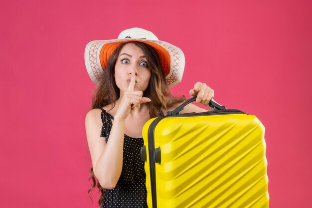 Menina linda em um vestido de bolinhas com chapéu de verão segurando a mala, olhando para a câmera, fazendo gesto de silêncio com o dedo nos lábios em pé sobre o fundo rosa