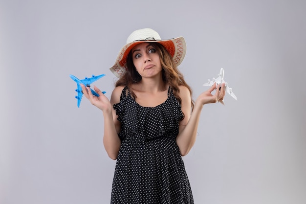 Menina linda em um vestido de bolinhas com chapéu de verão segurando aviões de brinquedo sem noção e confusa, olhando para a câmera em pé com os braços levantados, sem resposta, sobre fundo branco