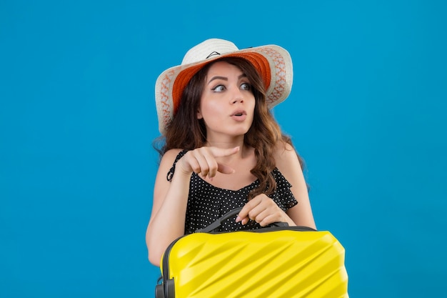 Menina linda em um vestido de bolinhas com chapéu de verão segurando uma mala e apontando com o dedo para o lado, parecendo surpresa em pé sobre um fundo azul