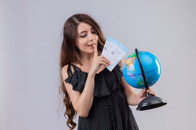 Menina linda em um vestido de bolinhas segurando passagens aéreas e um globo olhando para ele intrigada e feliz em pé sobre um fundo branco