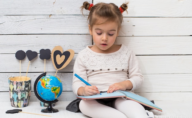 Menina linda garota da escola sentada sobre um fundo branco de madeira com um globo nas mãos e um caderno, o conceito de conhecimento