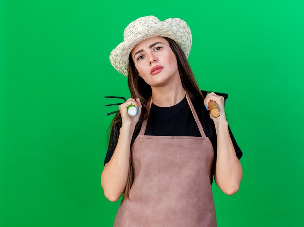 Menina linda jardineira desagradável de uniforme usando chapéu de jardinagem, colocando o ancinho com ancinho de enxada no ombro isolado no verde